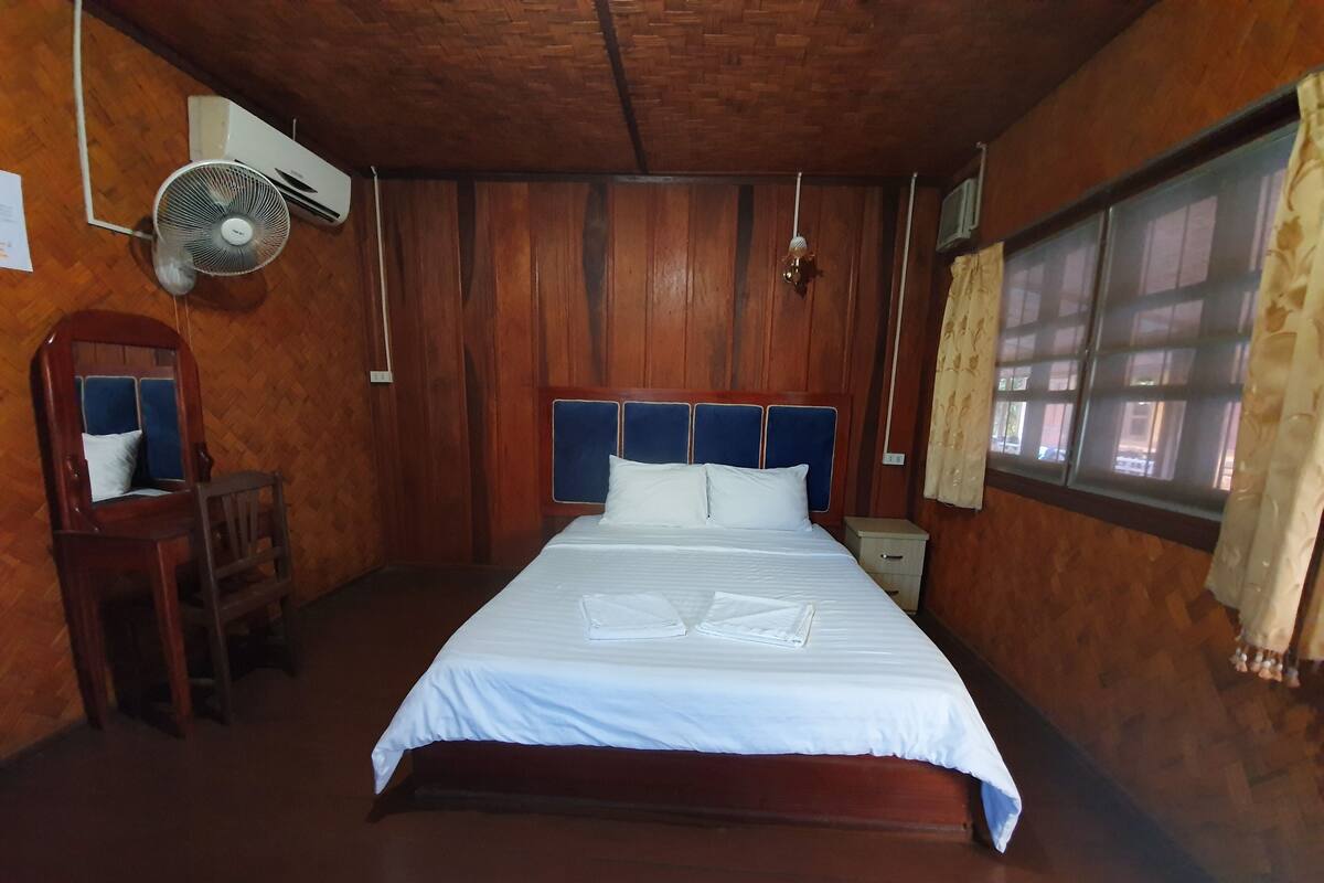 Een kamer met houten lambrisering, een bed, een stoel, een nachtkastje, een ventilator en een raam met gordijnen.