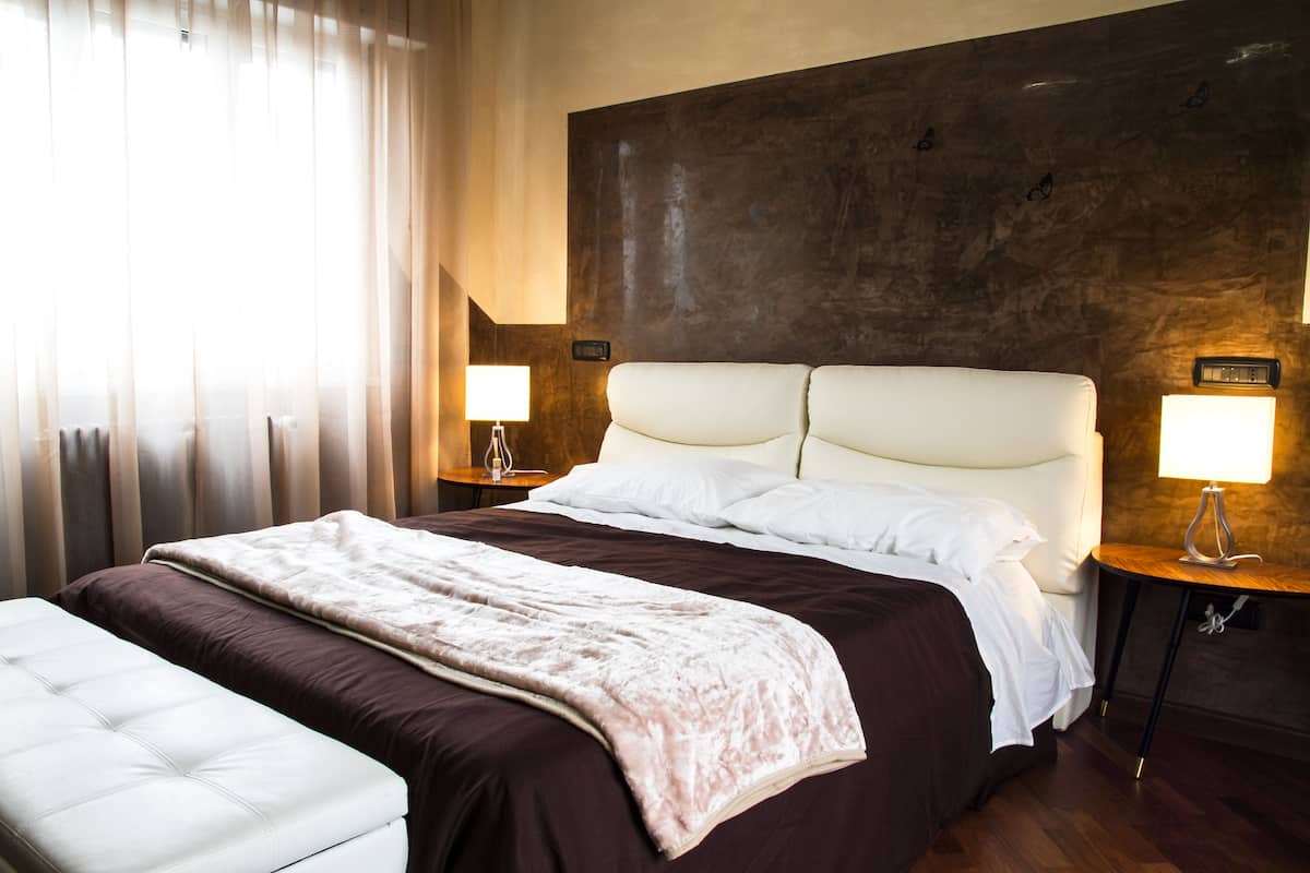 A hotel room with a large bed, two bedside tables with lamps, a headboard, and a window with curtains.
