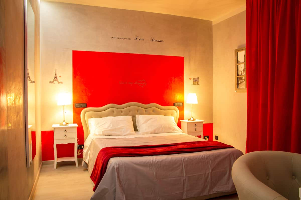 A hotel room with a bed, two bedside tables with lamps, a red curtain, and a wall with a red panel and text.