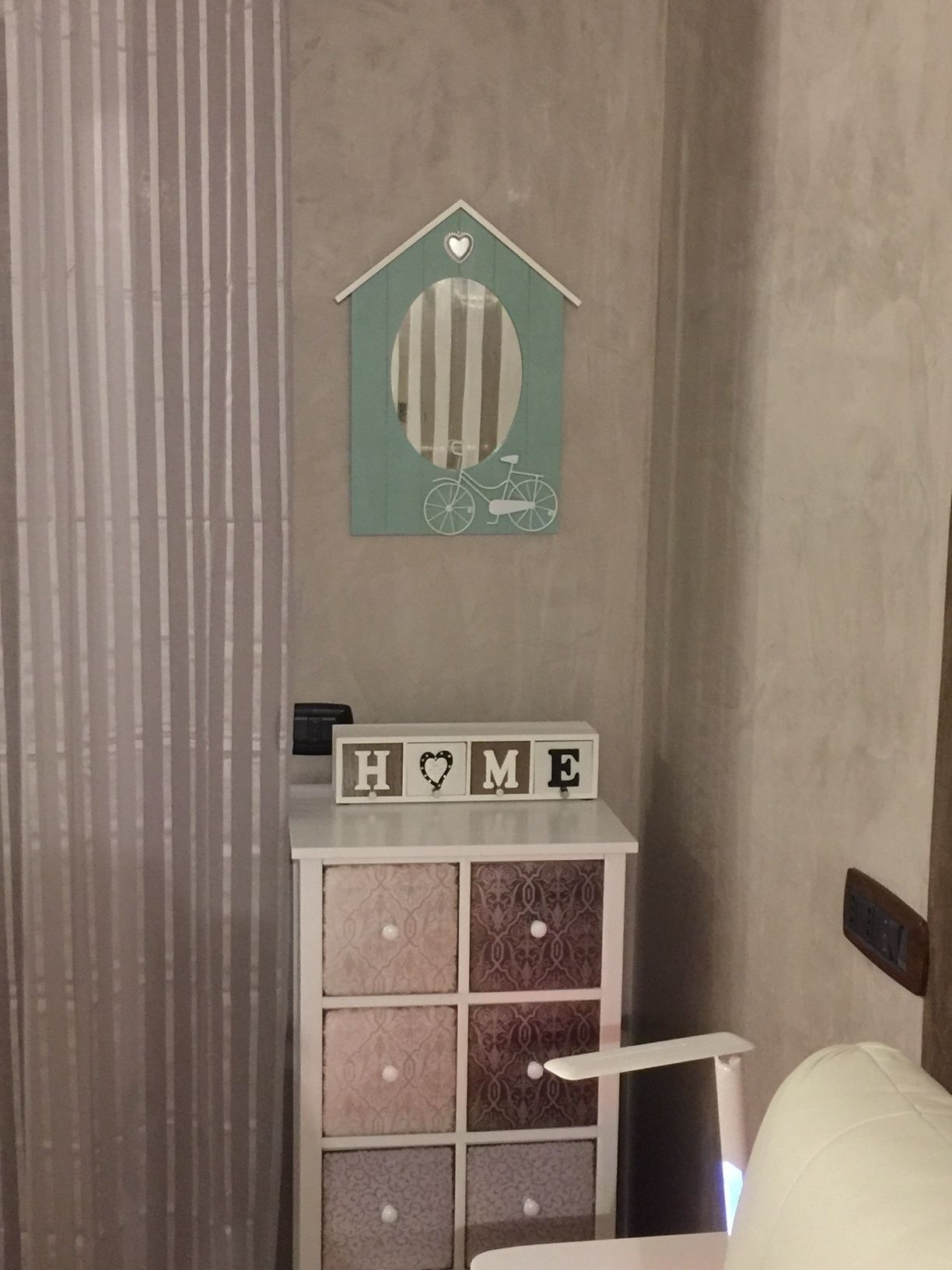 Ein Schlafzimmer mit einem Bett, einer Kommode mit Schubladen mit Muster sowie einer Wanddekoration eines Fahrrads und einem 'HOME'-Schild auf der Kommode.
