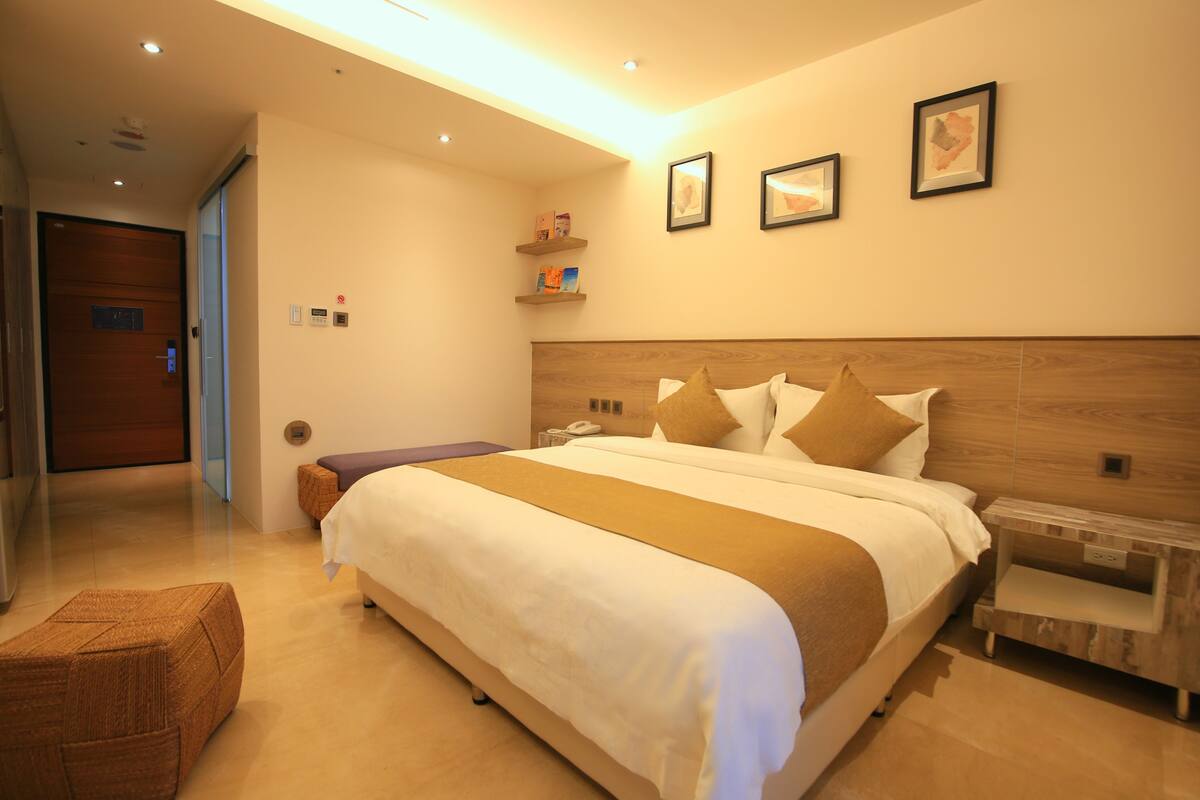 A modern hotel room with a large bed, a wooden headboard, a bedside table, and a small seating area.