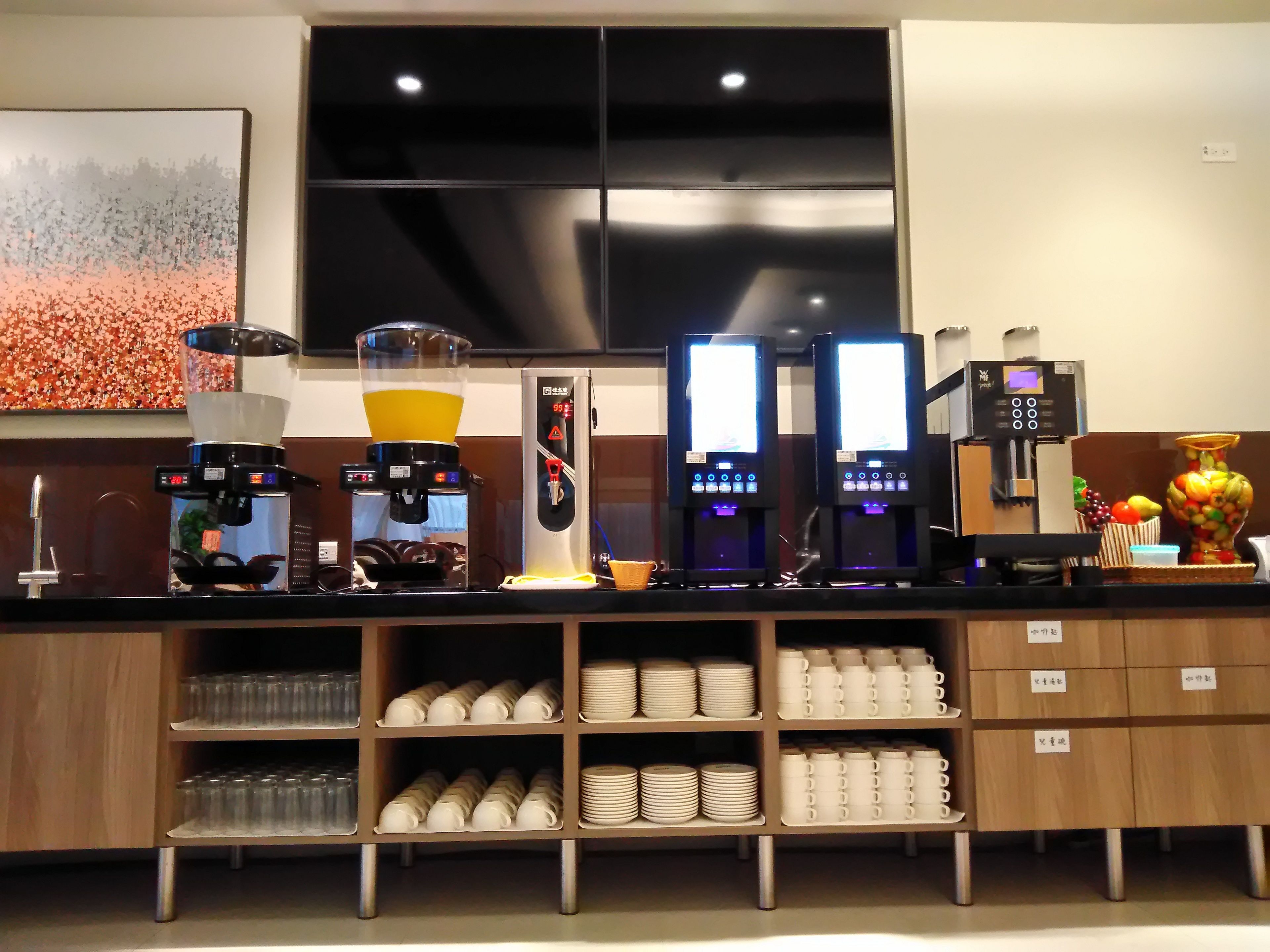 A coffee station with multiple coffee machines, a fruit display, and neatly arranged cups and plates on shelves.