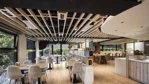 A modern dining area with wooden tables and chairs, a central bookshelf, and a view of greenery outside. - Dar Lon Hotel (Hsinchu)