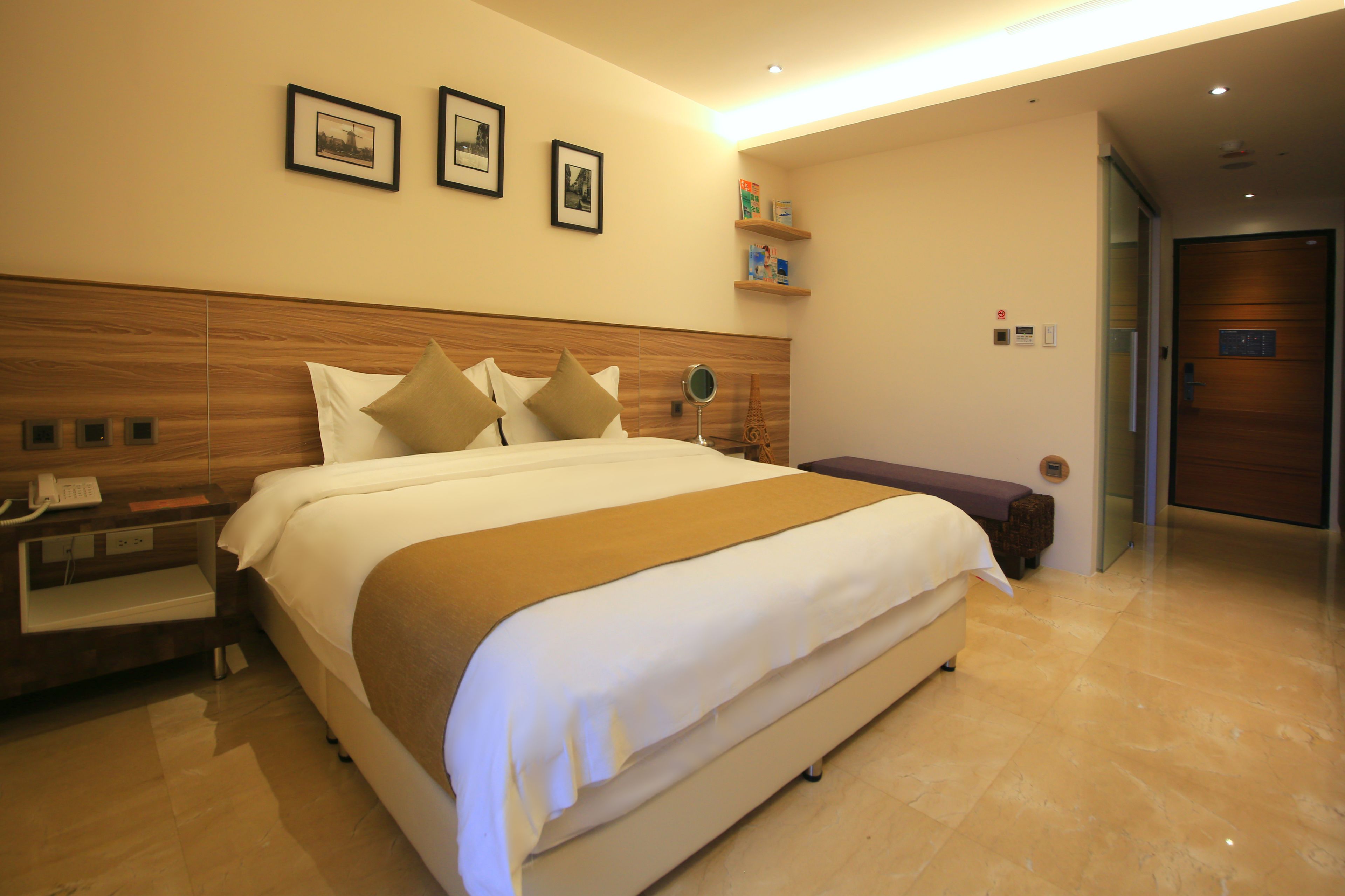 A hotel room with a large bed, wooden headboard, bedside table with a lamp, and a small shelf with toiletries.