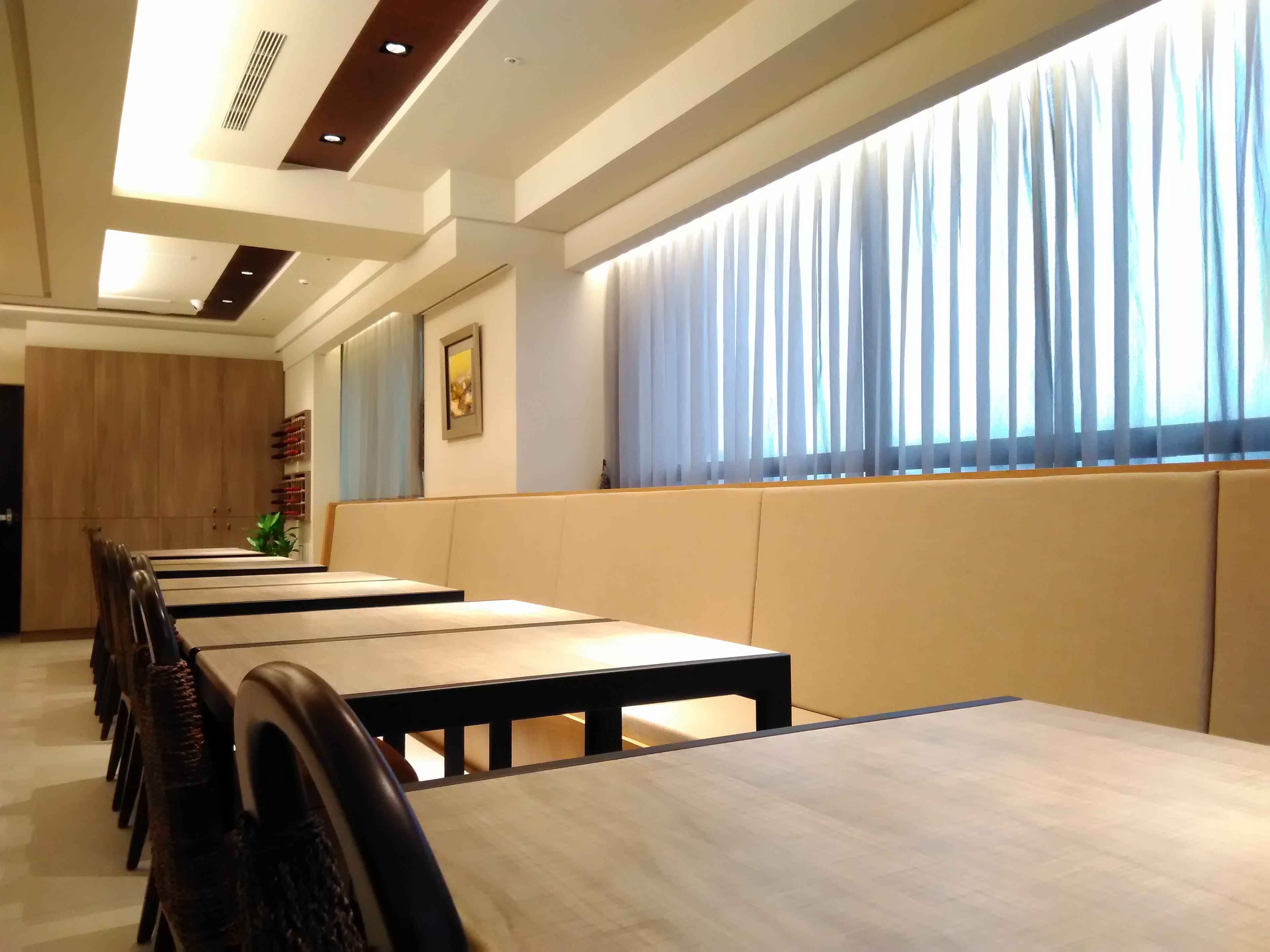 Un comedor con mesas y sillas largas, un panel de pared de madera y un amplio ventanal con cortinas.