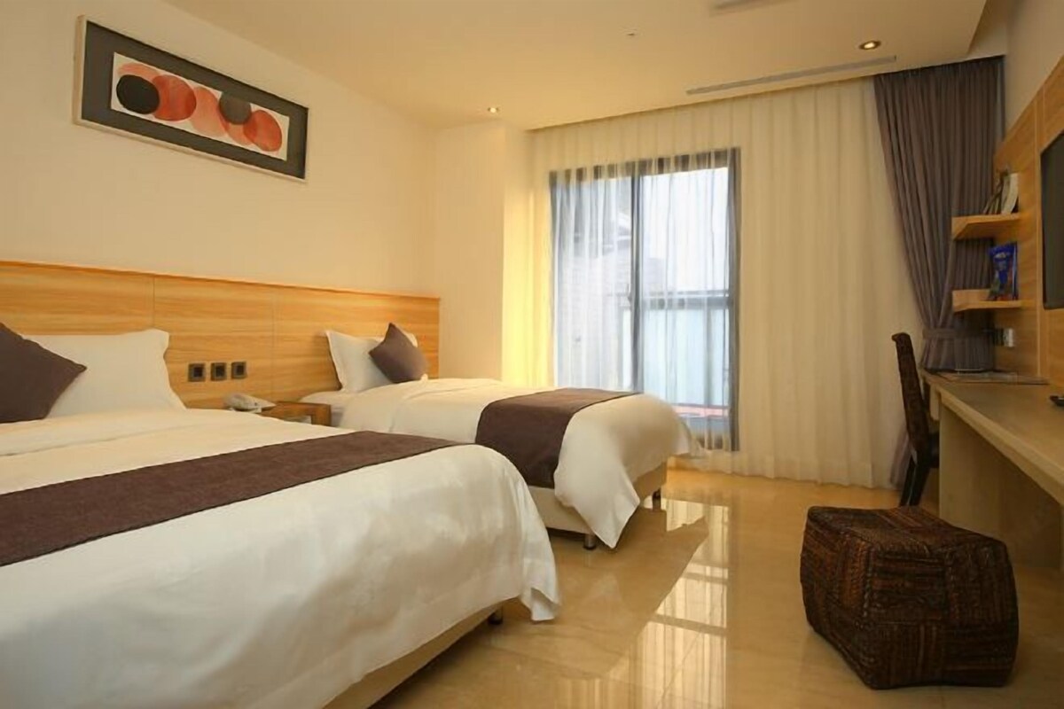 A hotel room with two beds, a wooden headboard, a large window with curtains, a small table, and a woven ottoman.