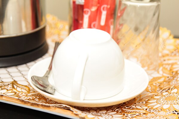 A white teapot on a white saucer with a spoon on a patterned tablecloth. - Streets of Goldbw B&B (Gaborone)