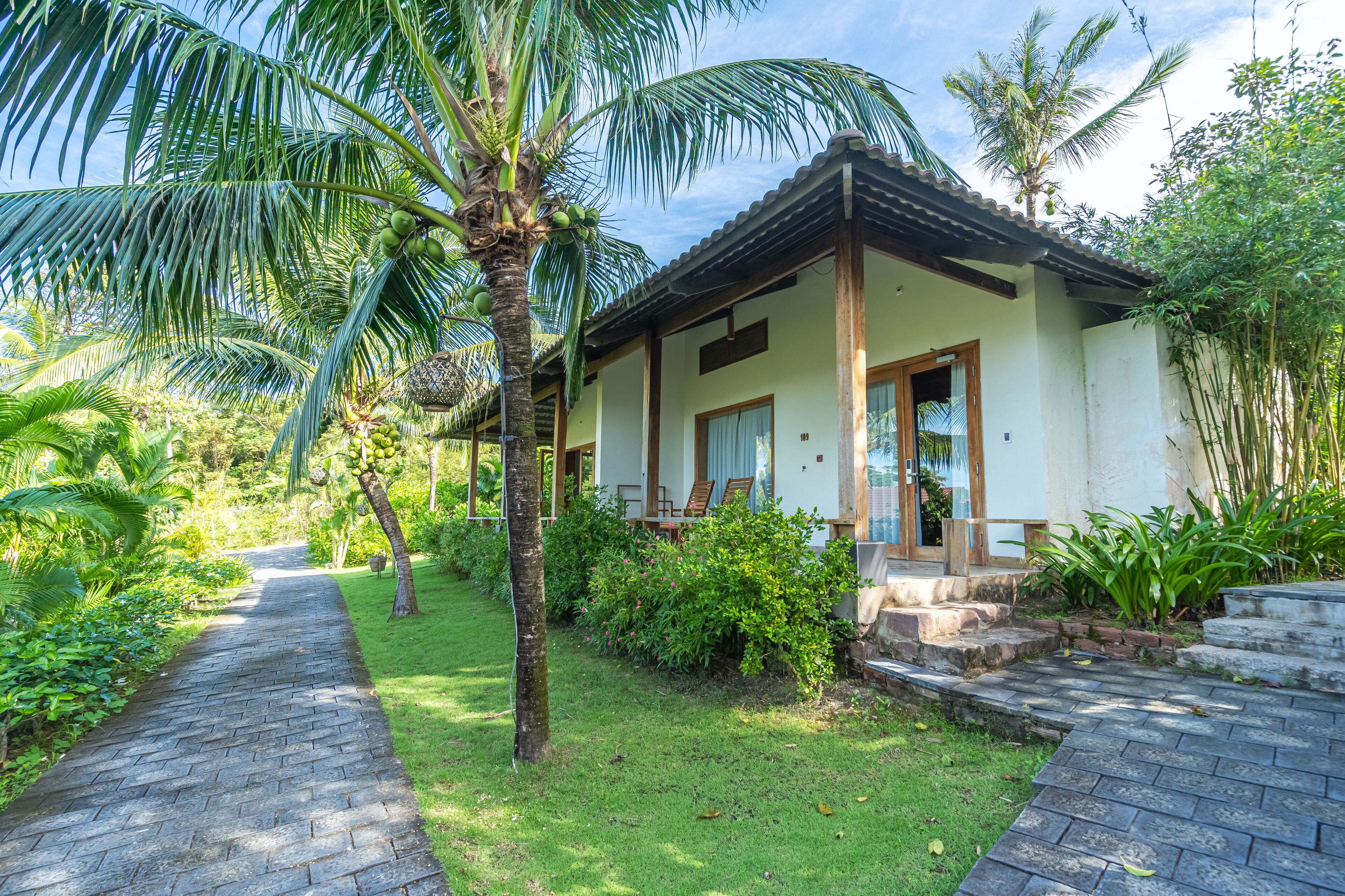 A bungalow with a paved pathway leading to the entrance, surrounded by lush greenery and palm trees.