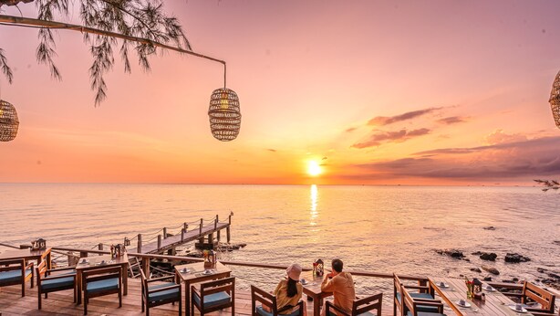 A wooden deck with tables and chairs facing a sunset over the water. - Camia Resort & Spa (Phu Quoc)