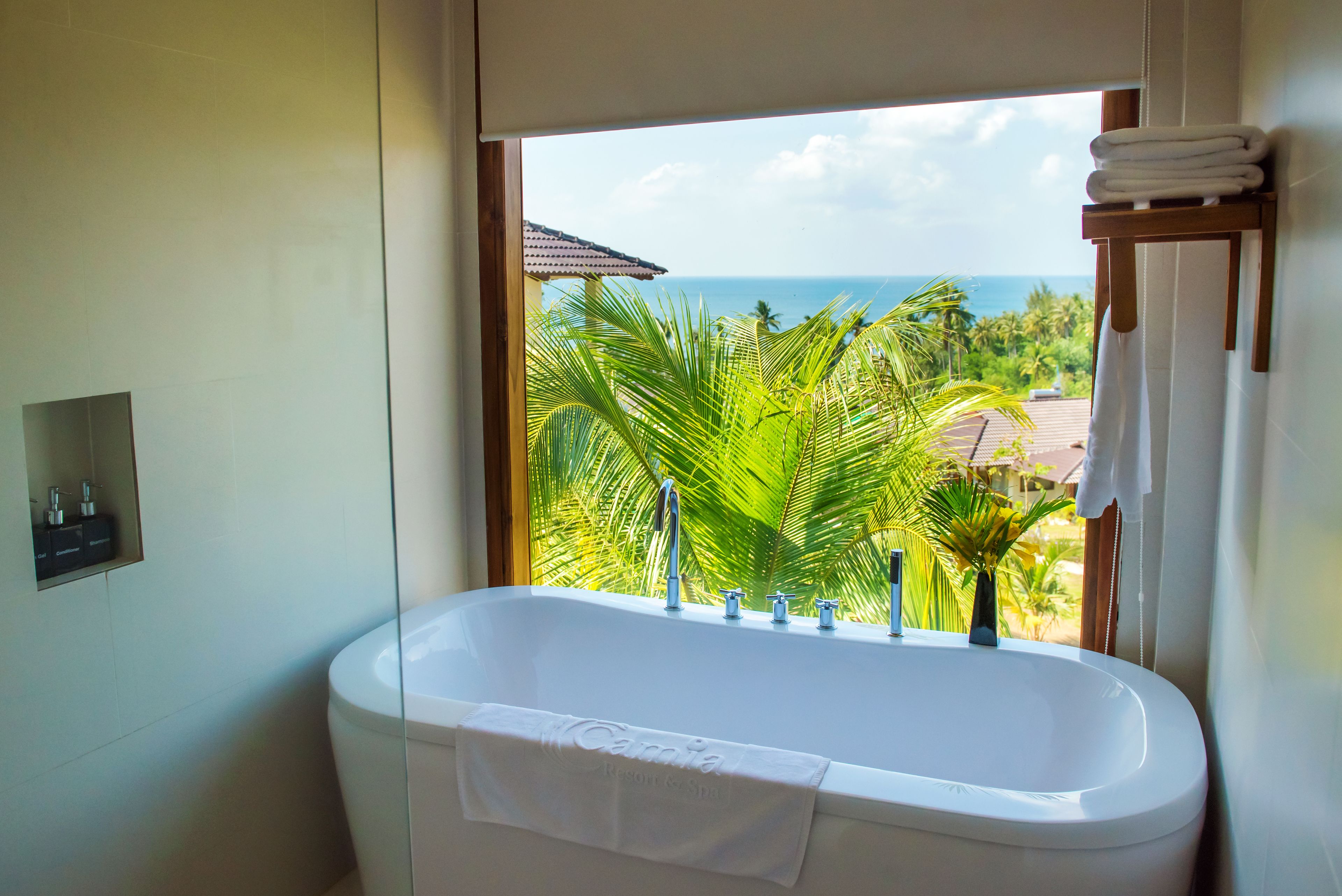 Une salle de bain avec une baignoire, une fenêtre donnant sur l’océan et les palmiers, et un porte-serviettes avec des serviettes pliées.