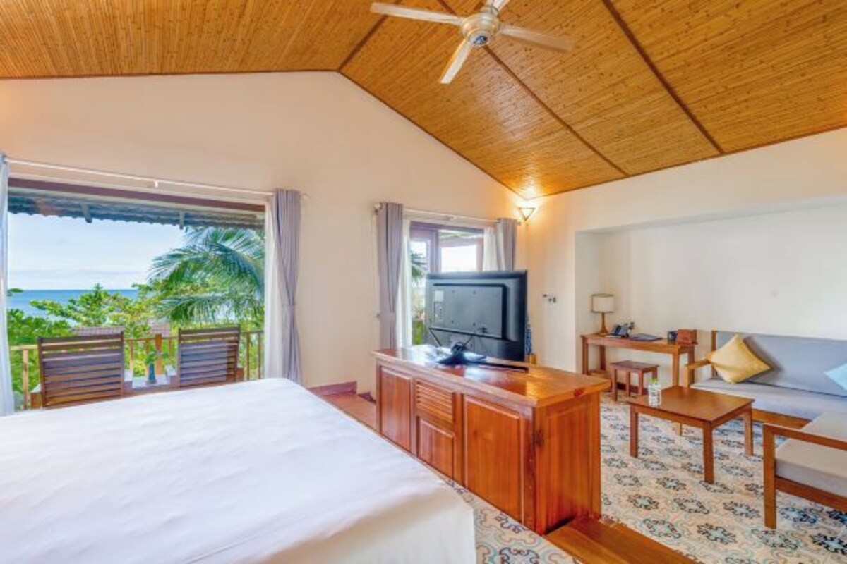 A hotel room with a bed, a TV, a desk, and a sofa. There is a balcony with a view of the ocean and palm trees.