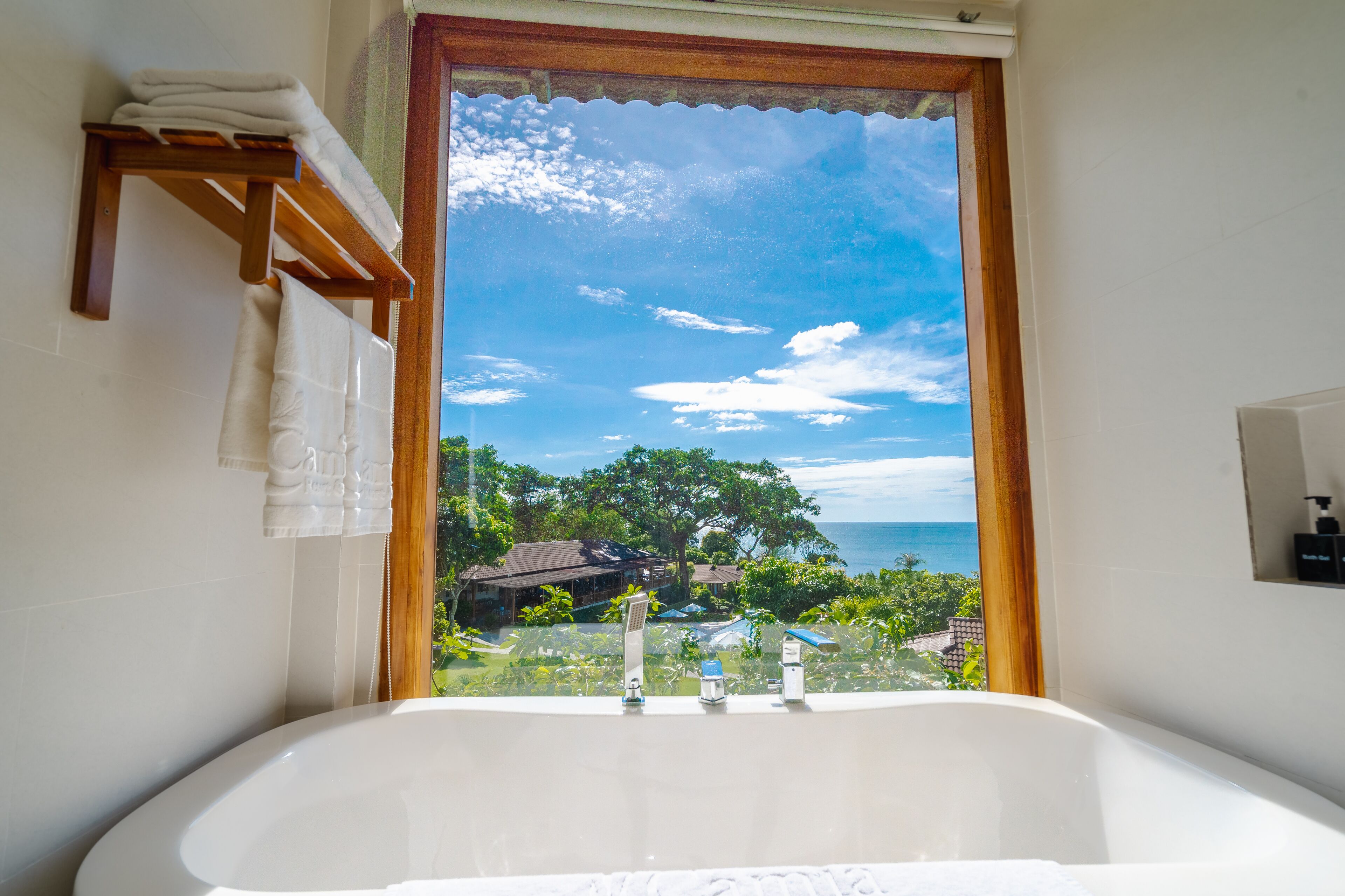 A bathroom with a large bathtub, a window with a view of the ocean and greenery, and a towel rack with folded towels.