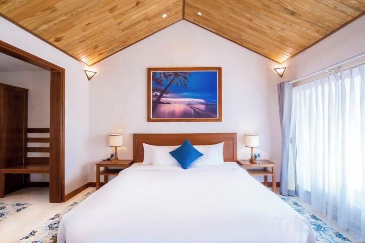 A hotel room with a large bed, wooden ceiling, a painting of a beach, and a window with curtains.