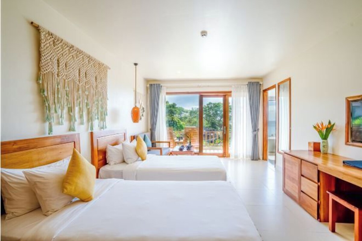 A hotel room with two beds, a wooden desk, and a large window offering a view of greenery.