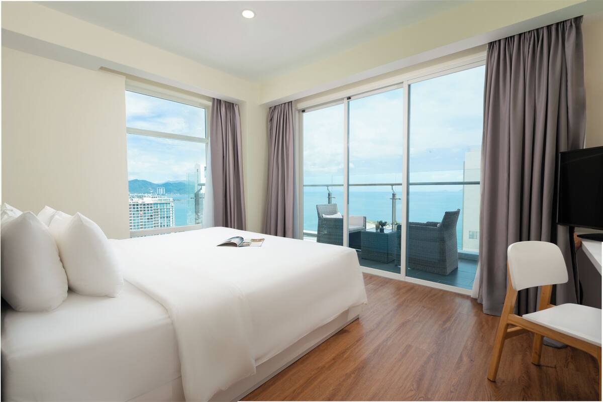 Ett hotellrum med en stor säng, en TV, en stol och en balkong med utsikt över havet och byggnaderna.