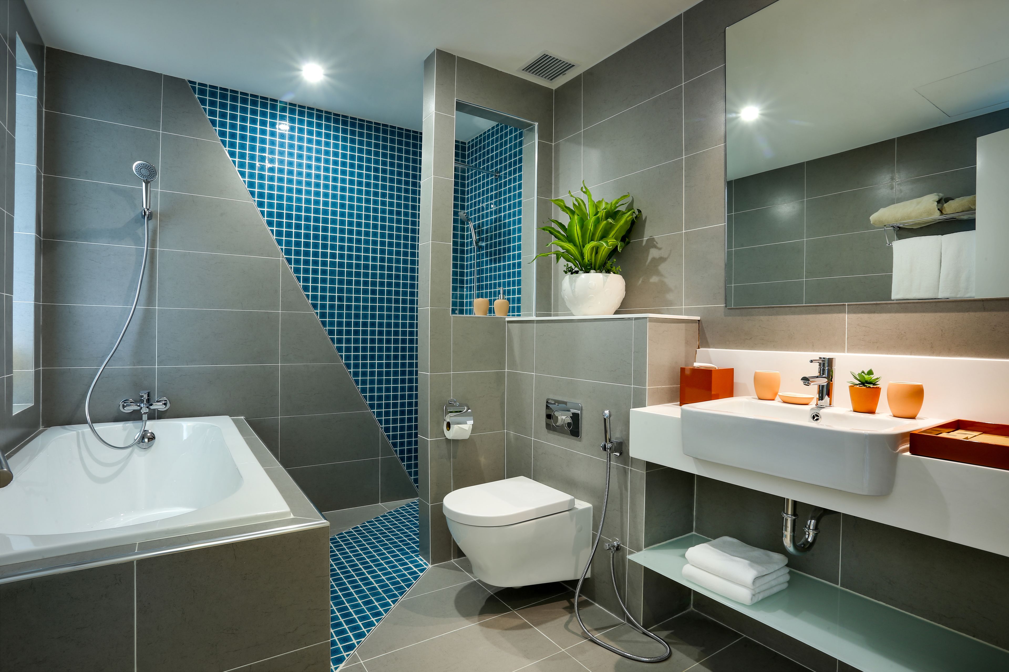 A modern bathroom with a bathtub, toilet, and sink, featuring blue and grey tiles, a potted plant, and a mirror.