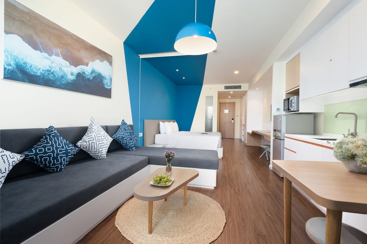 Ett modernt hotellrum med en grå soffa, ett träbord med en vas med blommor, en stor abstrakt målning, en blå hänglampa och ett litet kök i bakgrunden.