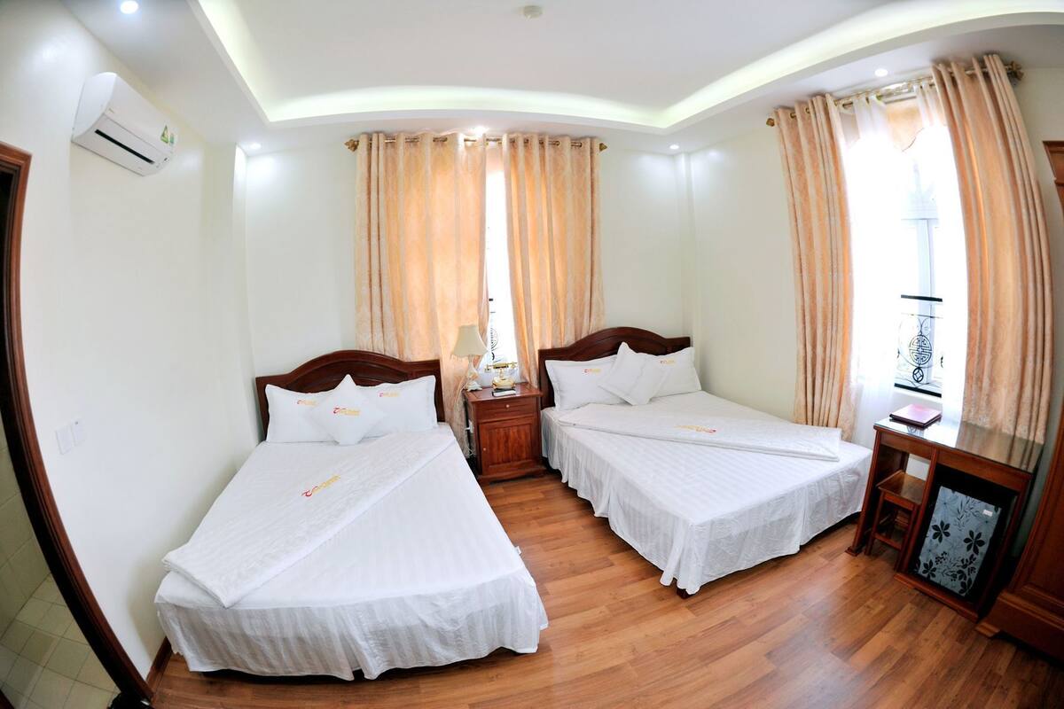 A hotel room with two beds, a wooden floor, an air conditioning unit, and a window with curtains.