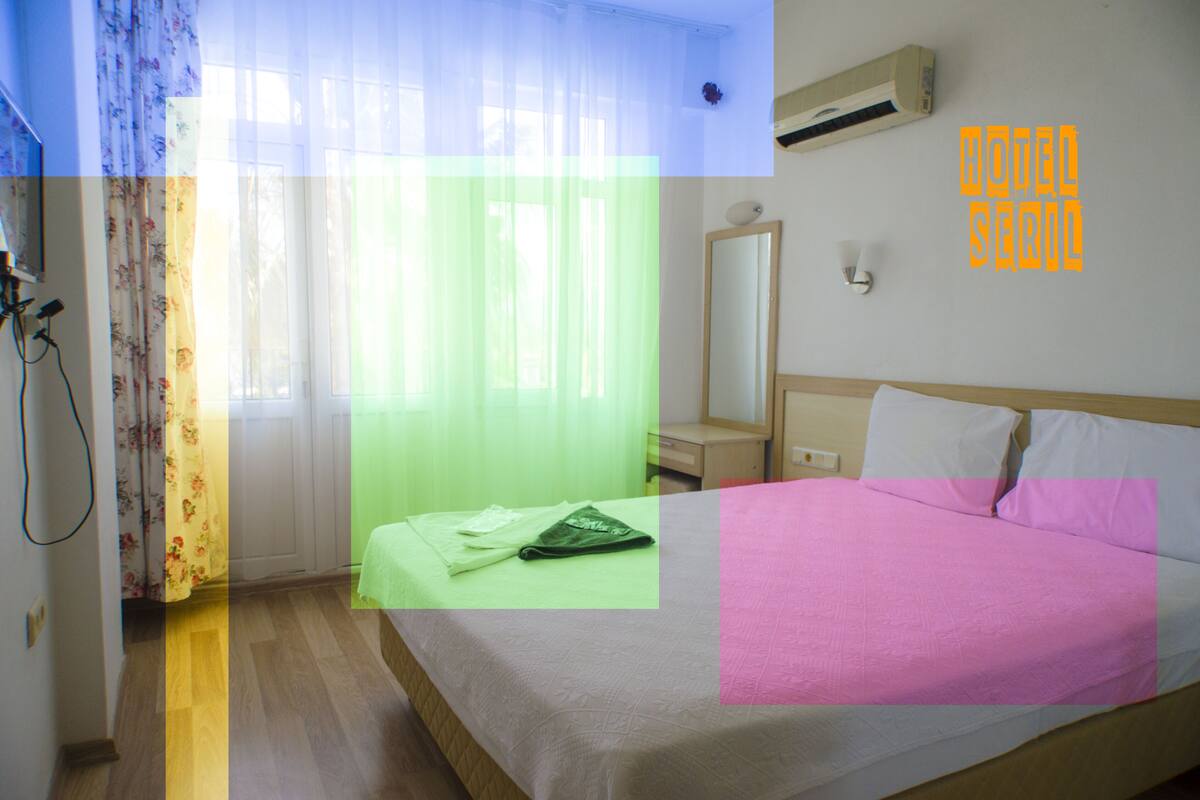 Quarto de hotel com cama, escrivaninha, espelho e janela com cortinas translúcidas.