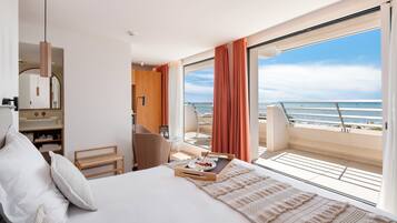 Une chambre d’hôtel avec un lit, un bureau, une chaise, une petite table sur laquelle se trouve un plateau de nourriture, et une vue sur la mer.