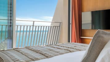 Une chambre d’hôtel avec un balcon donnant sur la mer, un lit recouvert d’une couverture à motifs, et un téléviseur fixé au mur.