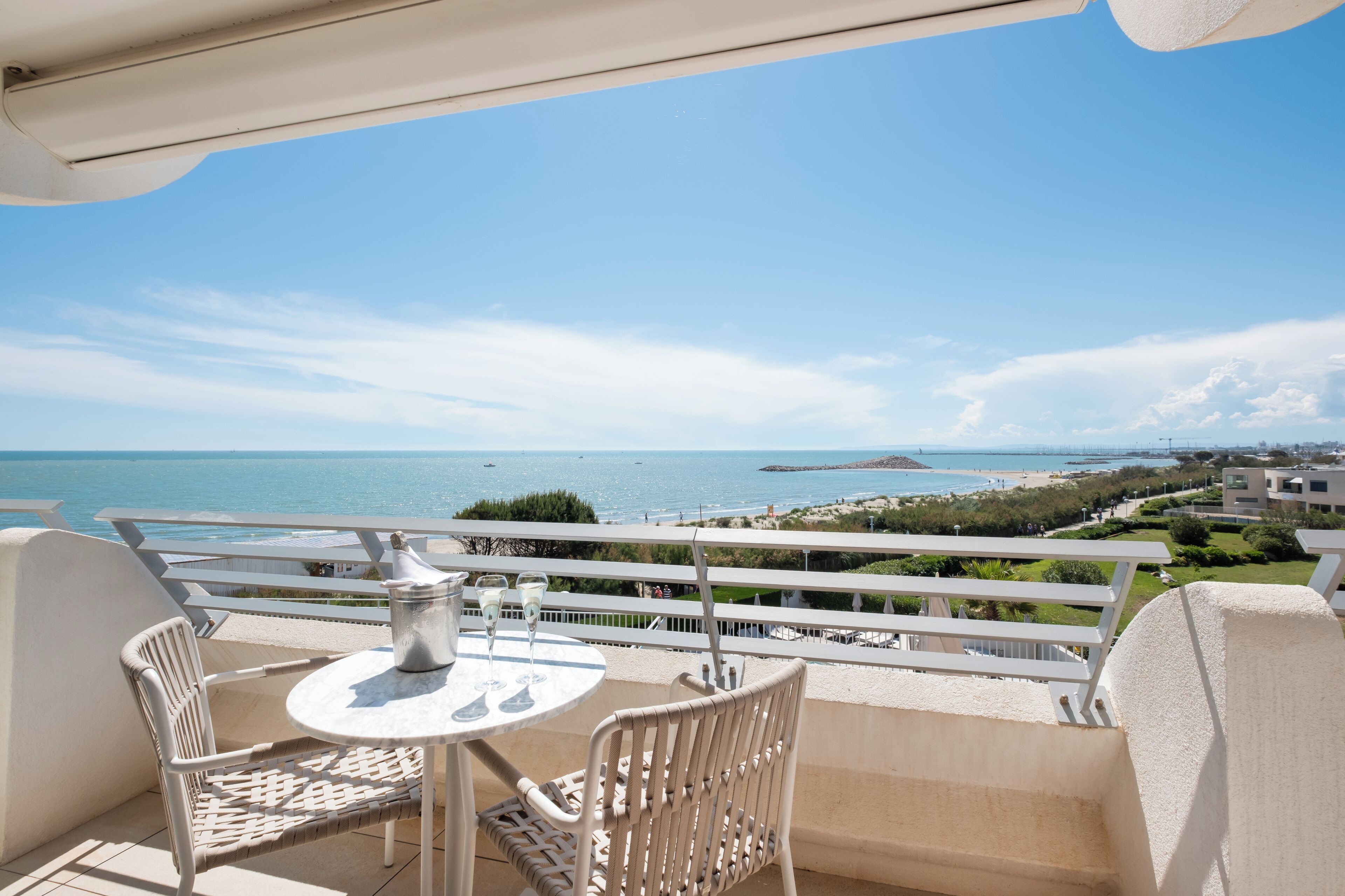 A balcony with a table and chairs, overlooking a coastal view with buildings and greenery.
