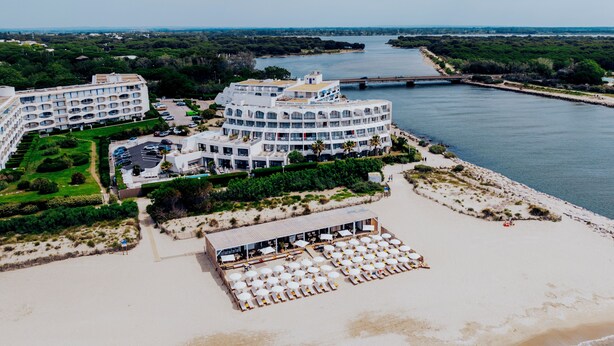 Un hotel frente al mar con varias edificaciones, una zona de playa con tumbonas y un río al fondo.