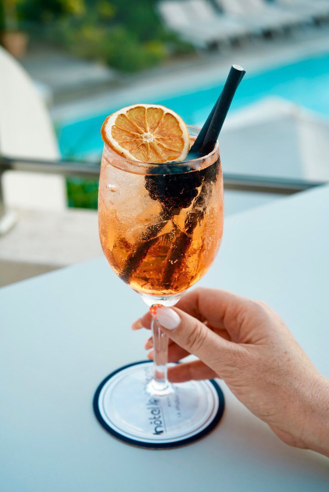 A hand holding a cocktail glass with a slice of orange and a straw, placed on a table with a pool in the background.