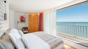 Un dormitorio con una cama grande, una mesita de noche, un armario de madera, un cuadro enmarcado en la pared y un balcón con vista al mar.