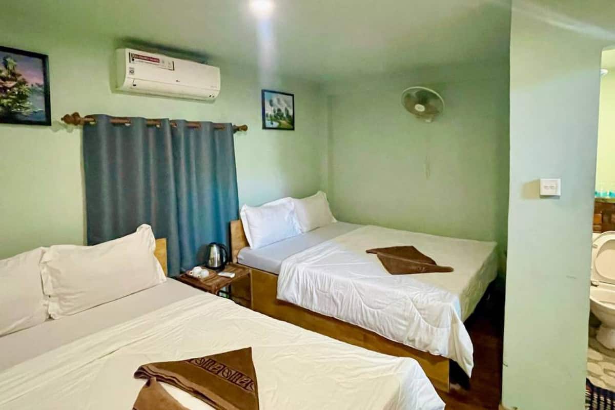 A hotel room with two single beds, a small table with a kettle, a wall-mounted air conditioner, and a ceiling fan.