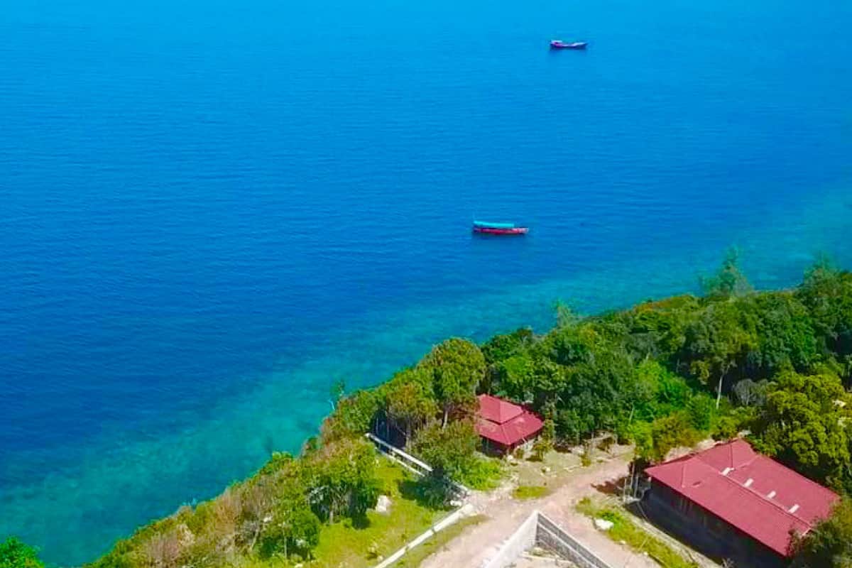 An aerial view of a coastal area with two buildings, one with a red roof and another with a blue roof, surrounded by lush greenery and clear blue water.