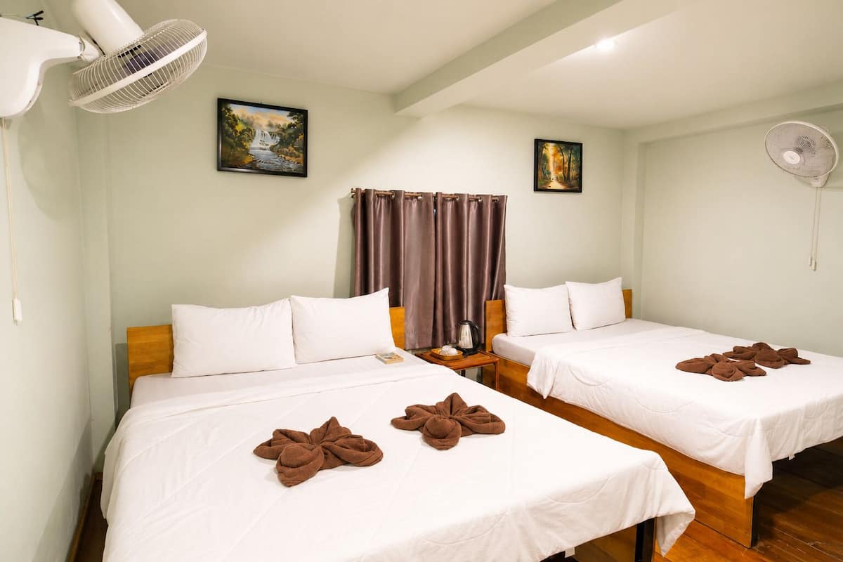 A hotel room with two beds, a window with curtains, a painting on the wall, and a fan.