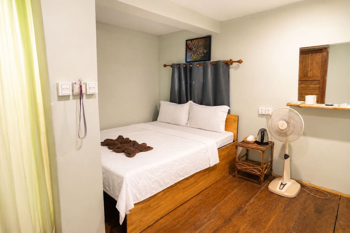 A bedroom with a bed, a bedside table with a fan, a wooden nightstand, a wall-mounted shelf, and a window with curtains.