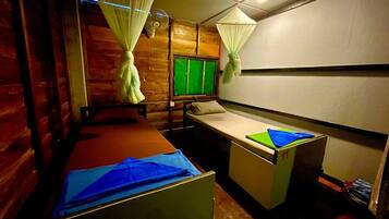 A small, compact room with two beds, a window with green blinds, and a fan.