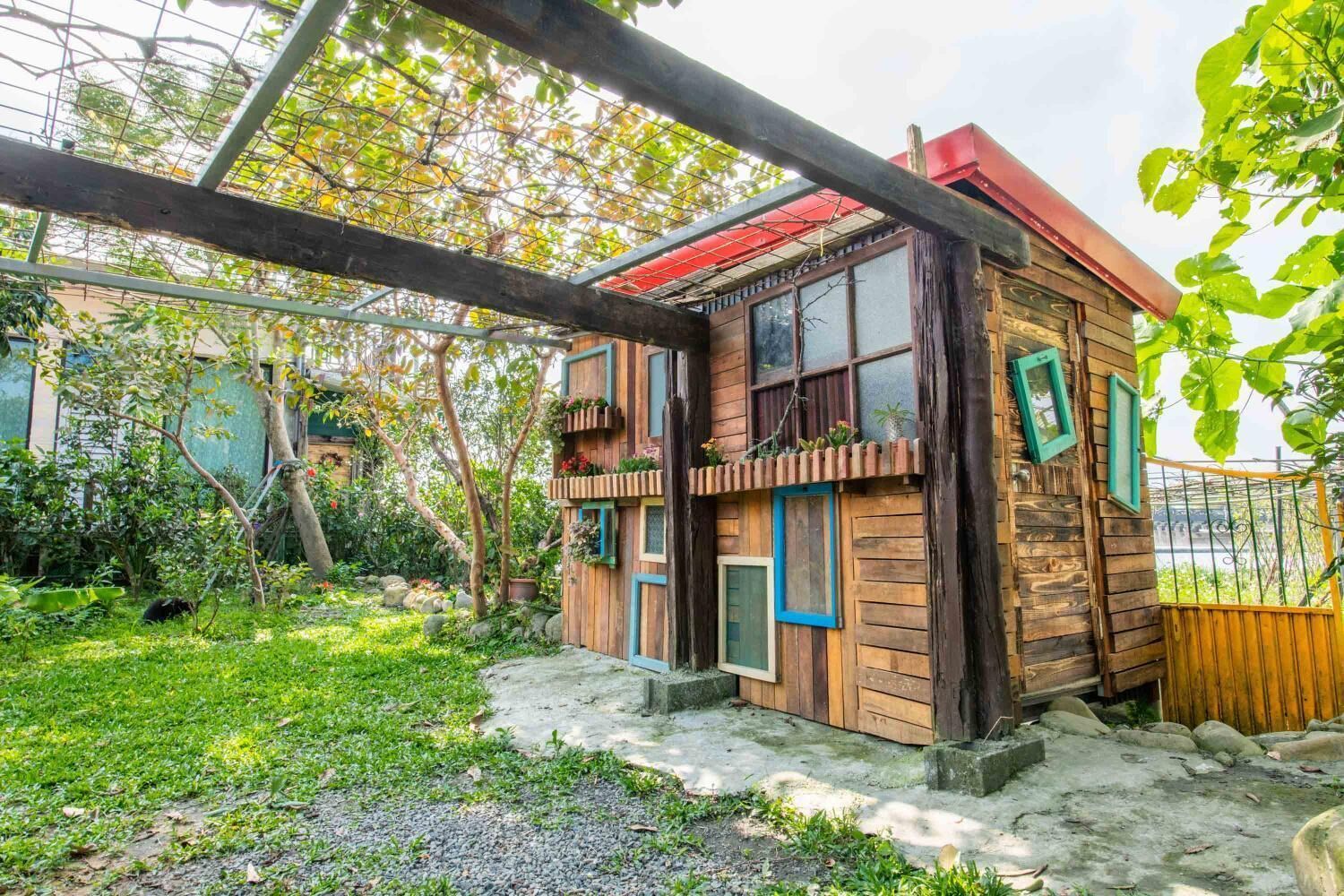 Une maison de campagne en bois, avec un toit rouge, entourée d’un jardin où poussent différentes plantes et arbres.