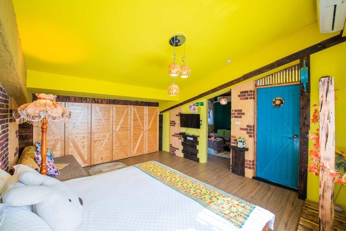 A bright yellow room with a bed, a wooden wardrobe, a blue door, and a wooden floor.