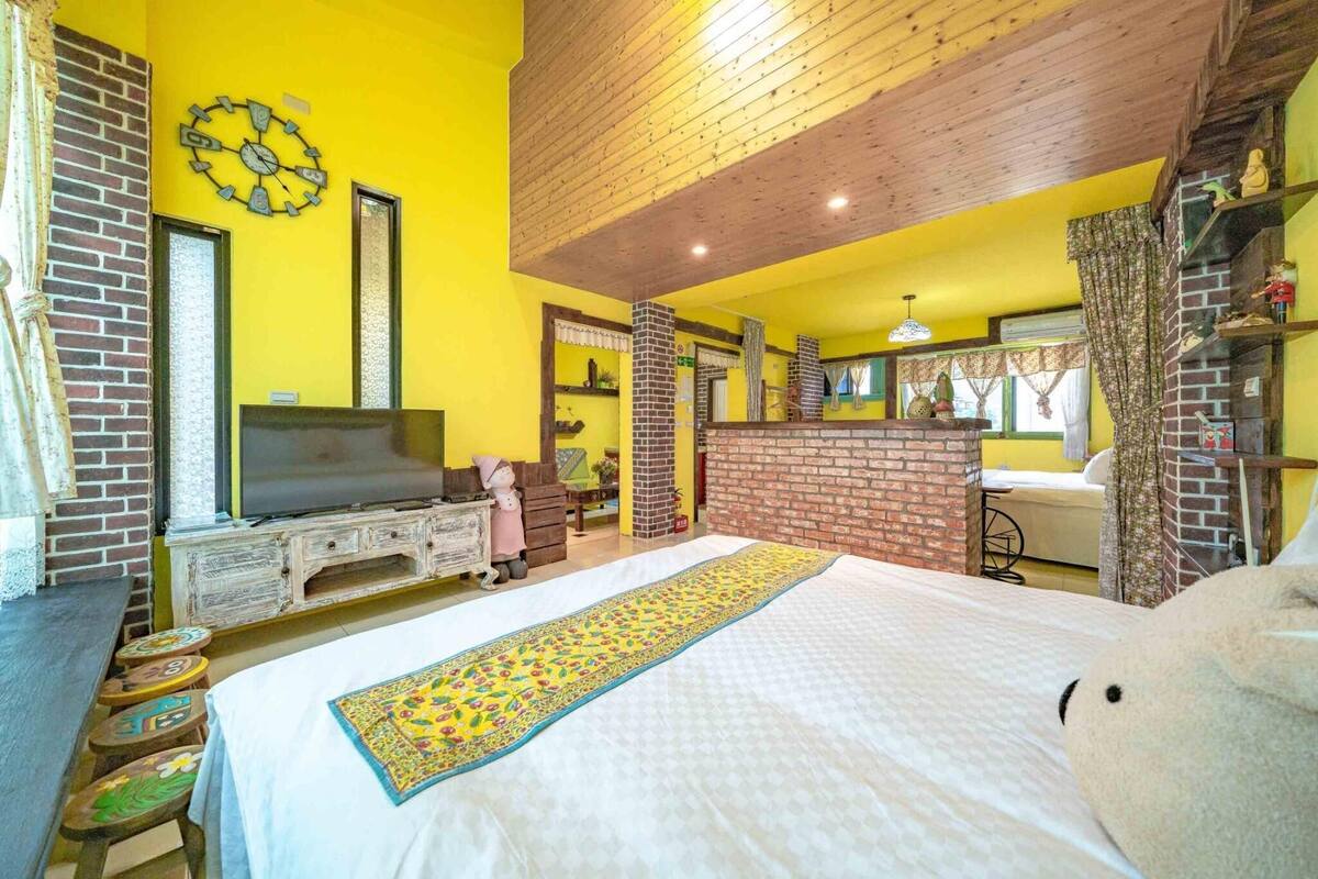 A room with a bed, a television, a brick fireplace, and a yellow wall.