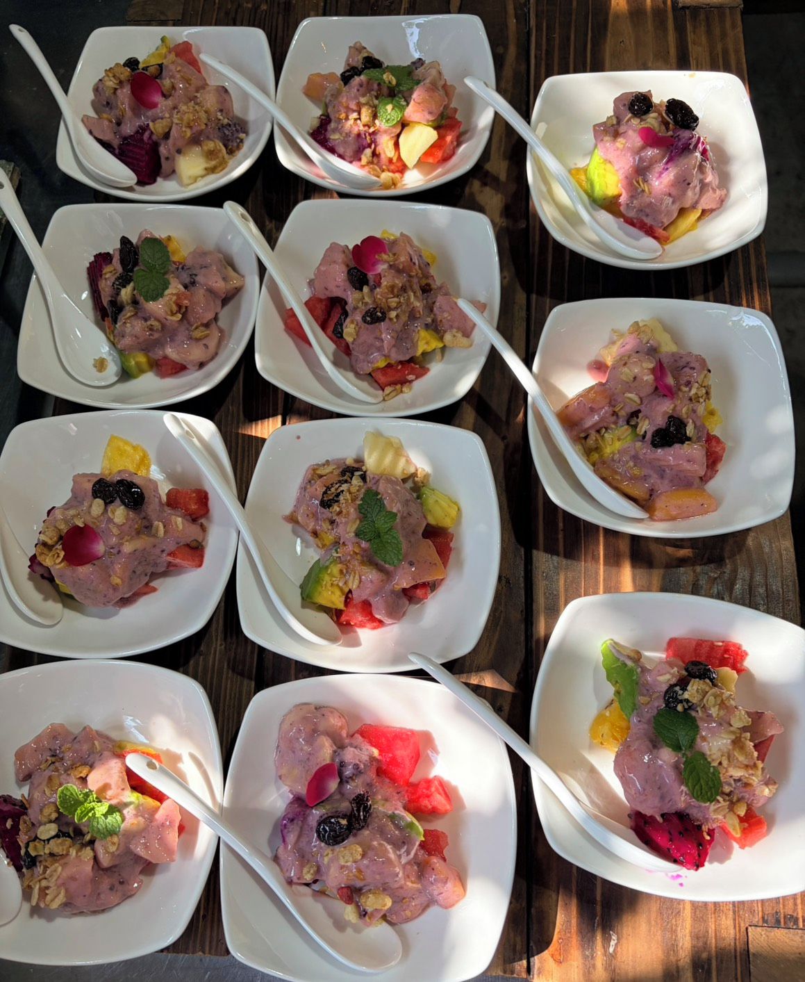 A series of square white plates with food, each accompanied by a white plastic spoon.