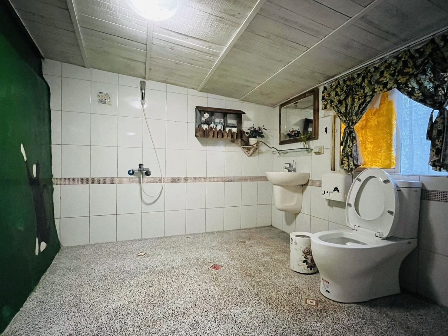 A bathroom with a white sink, toilet, and shower. There is a mirror above the sink and a window with a curtain.
