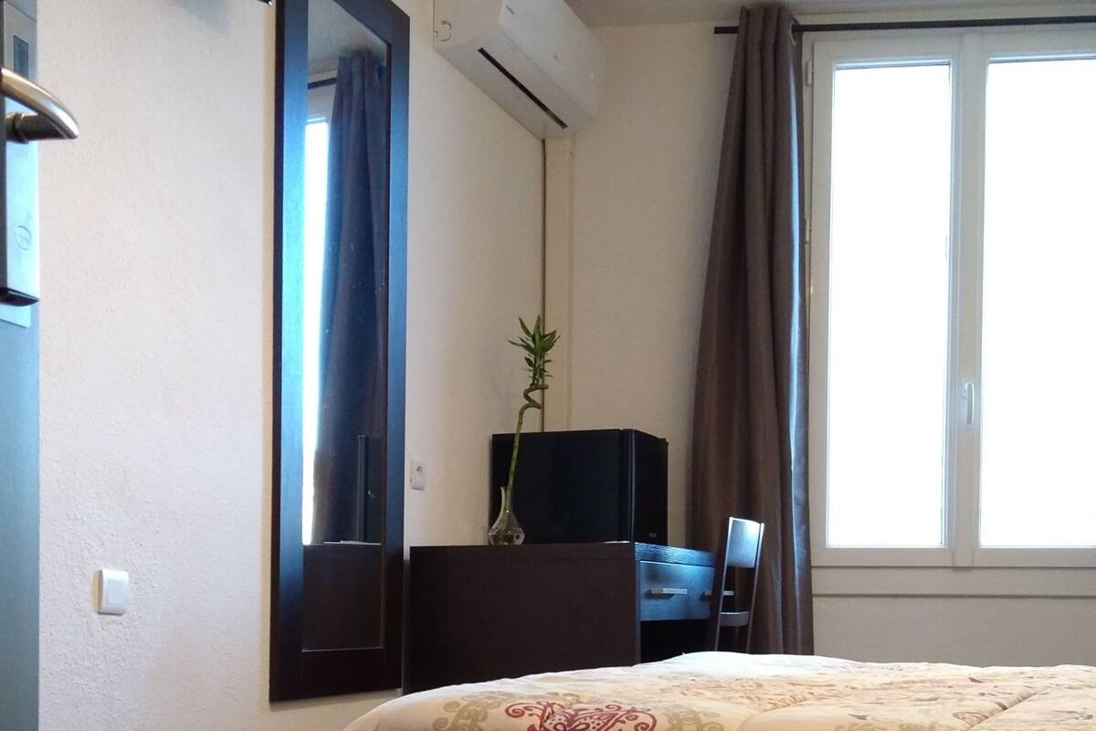 Habitación de hotel con cama, televisor de pantalla plana montado en la pared, unidad de aire acondicionado, escritorio con silla y ventana con cortinas.