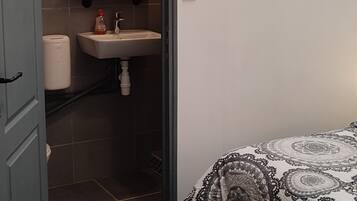 Une petite chambre à coucher, de dimensions réduites, comprenant un lit, une table de chevet, une étagère fixée au mur, et une salle de bain avec des toilettes, un lavabo et un porte-serviettes.