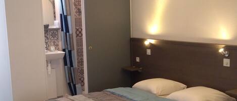 A hotel room with two beds, a bathroom with a shower curtain, a ceiling light fixture, and a wall-mounted light.