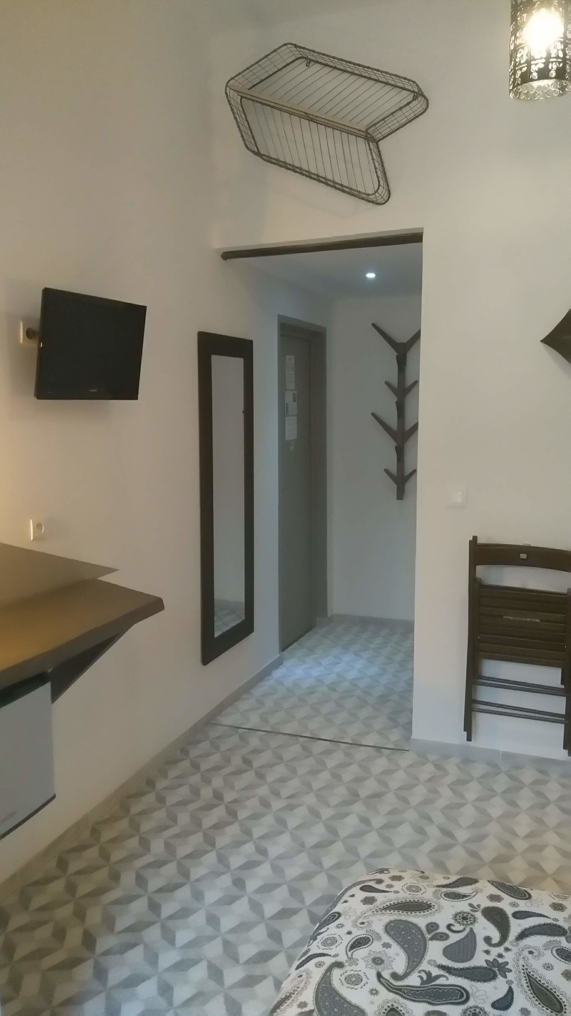 Una camera con un letto, una TV fissata al muro, uno specchio, una sedia di legno e un tappeto decorato.