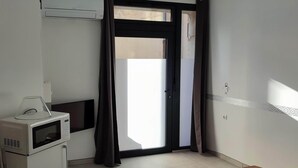 A narrow hallway with a microwave and a small refrigerator on the left, a sliding glass door to a balcony on the right, and a wall-mounted air conditioning unit above the door. - Mauguio Center Hôtel (Mauguio)