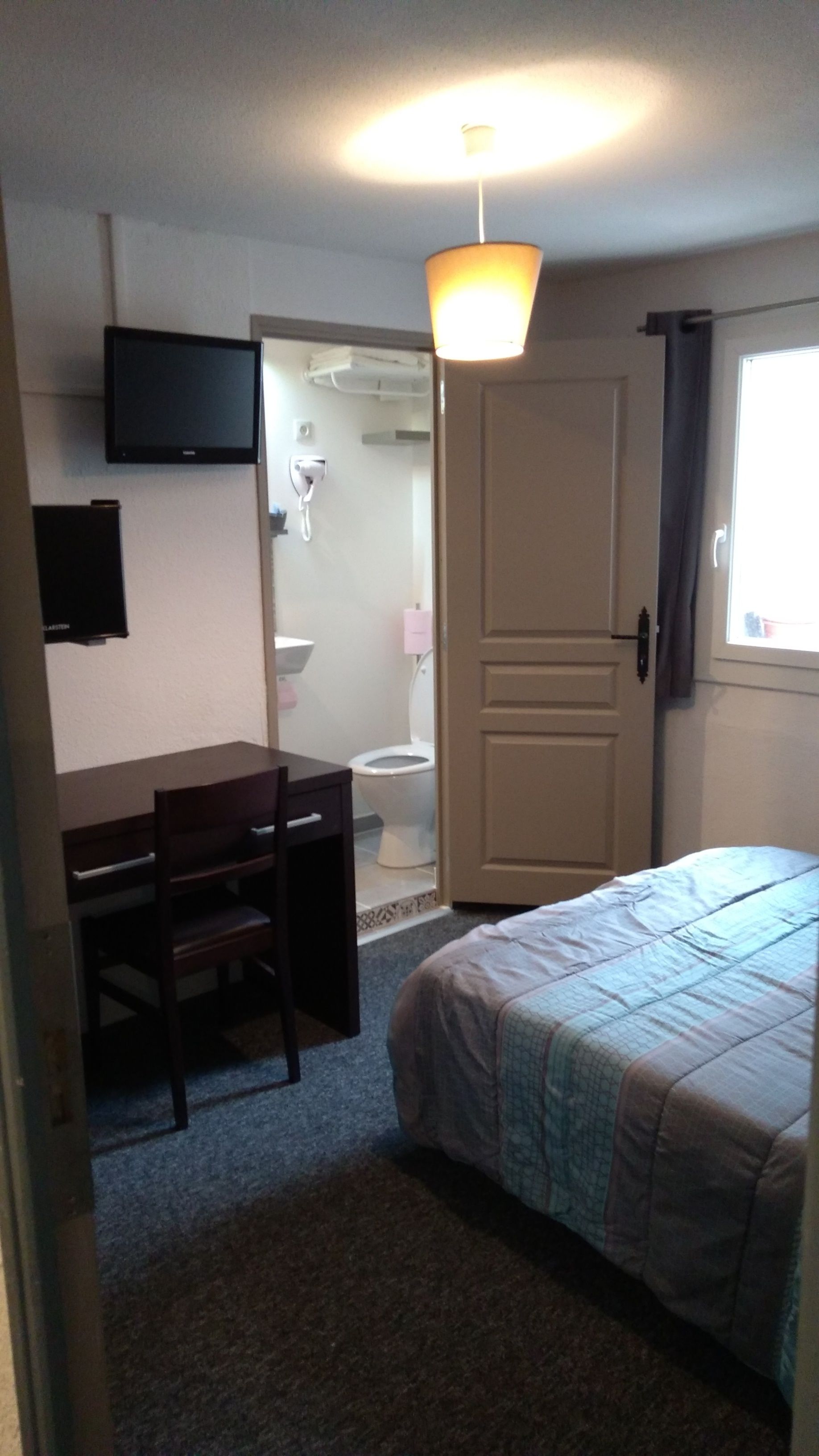 Ein Schlafzimmer mit Bett, Schreibtisch mit Stuhl, Fernseher an der Wand und einem durchgehenden Türspalte sichtbaren Badezimmer.