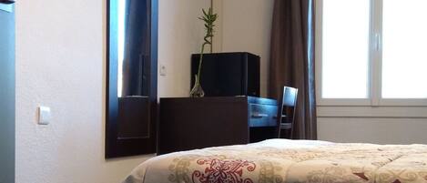 Habitación de hotel con cama, televisor de pantalla plana montado en la pared, escritorio con silla, planta en maceta y ventana con cortinas.