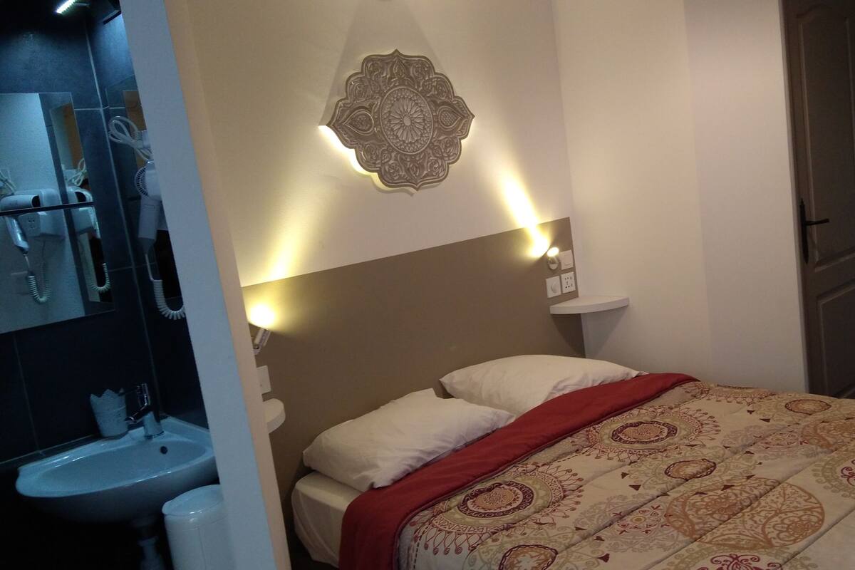 Habitación de hotel con cama, mesita de noche, espejo, lavabo y puerta.