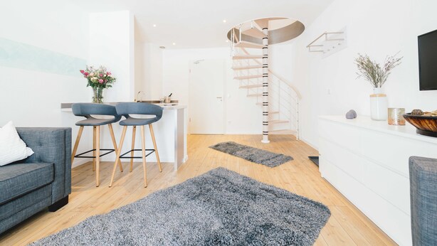 A modern living room with a grey sofa, two bar stools, a coffee table, a spiral staircase, and a white wall with a flat-screen TV.