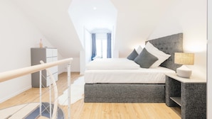 A modern bedroom with a sloped ceiling, a bed with grey bedding, a bedside table with a lamp, and a wardrobe.