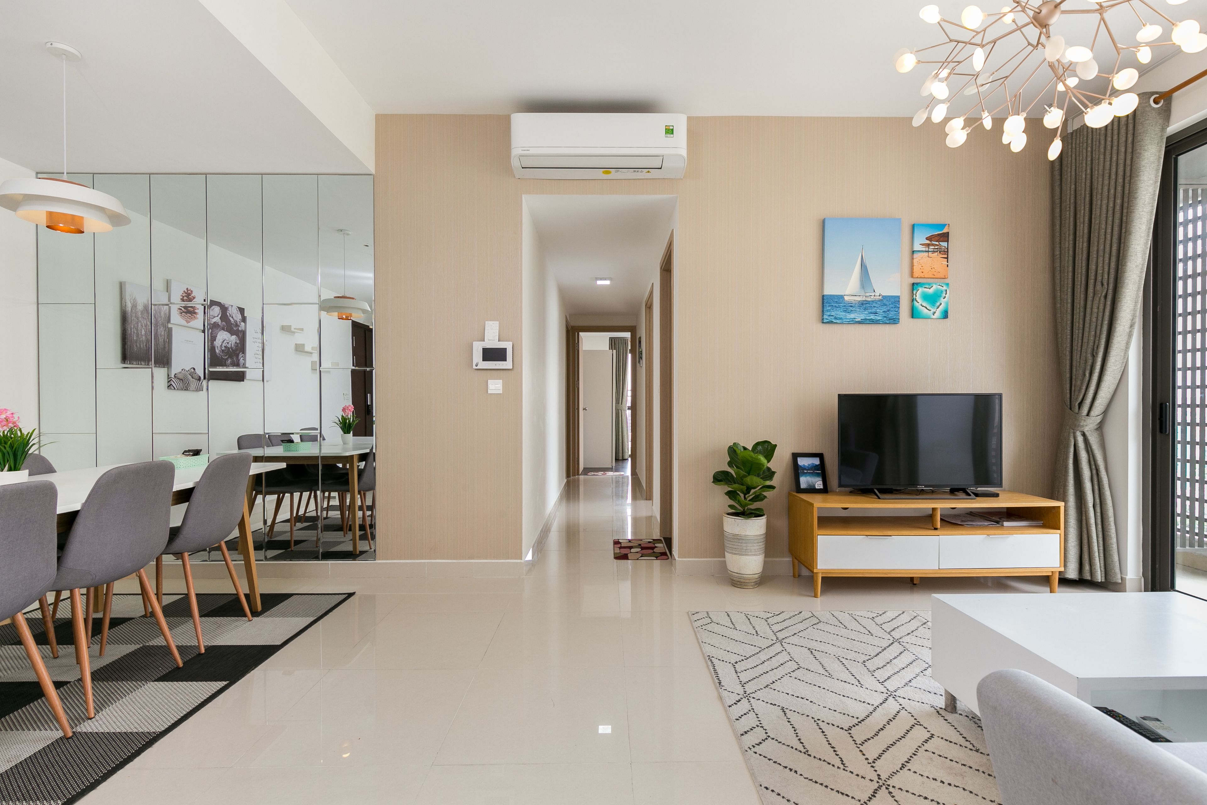 A modern living room with a flat-screen TV, a dining area with a glass partition, and a hallway leading to other rooms.